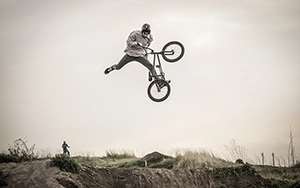 Ein einzelner BMX-Fahrer ist im oberen Teil eines großen Sprungs abgebildet, sein Fahrrad ist seitlich gedreht, während er hoch über dem Boden vor einem klaren, hellen Himmel schwebt.